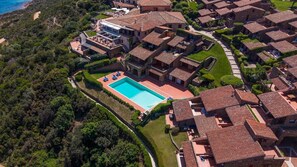 Vue aérienne d’un quartier résidentiel situé sur la côte, avec des maisons aux toits de tuiles rouges, une piscine centrale et une végétation luxuriante.