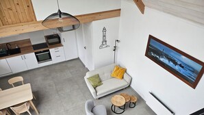 Une cuisine moderne avec des meubles blancs, un coin repas avec une table et des chaises en bois, un canapé gris avec des coussins et un tableau encadré au mur.