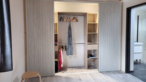 Un dressing accessible sans porte, avec des étagères en bois et une tringle pour suspendre les vêtements, une chaise, et une salle de bain partiellement visible avec un lavabo blanc.