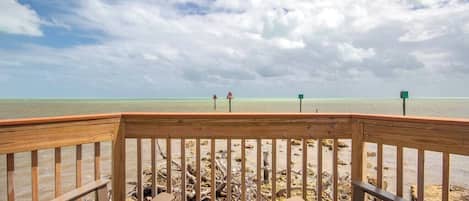 A wooden deck with benches overlooking a beach with a clear view of the ocean and a thatched roof overhead.