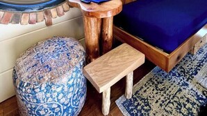 Una habitación acogedora con un ventanal redondo, una cama, una pequeña mesa de madera, un otomano estampado y un estante fijado a la pared.