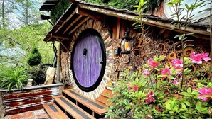 Un edificio rústico con una puerta morada, bancos de madera y una planta en maceta con flores rosadas.