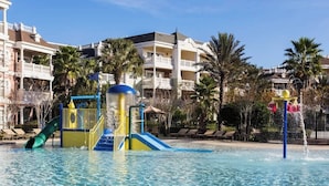A swimming pool with a slide and water play area, surrounded by palm trees and hotel buildings.
