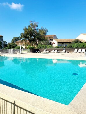 Une piscine d’un bleu éclatant, bordée de chaises longues blanches, avec une clôture blanche et des bâtiments en arrière-plan.