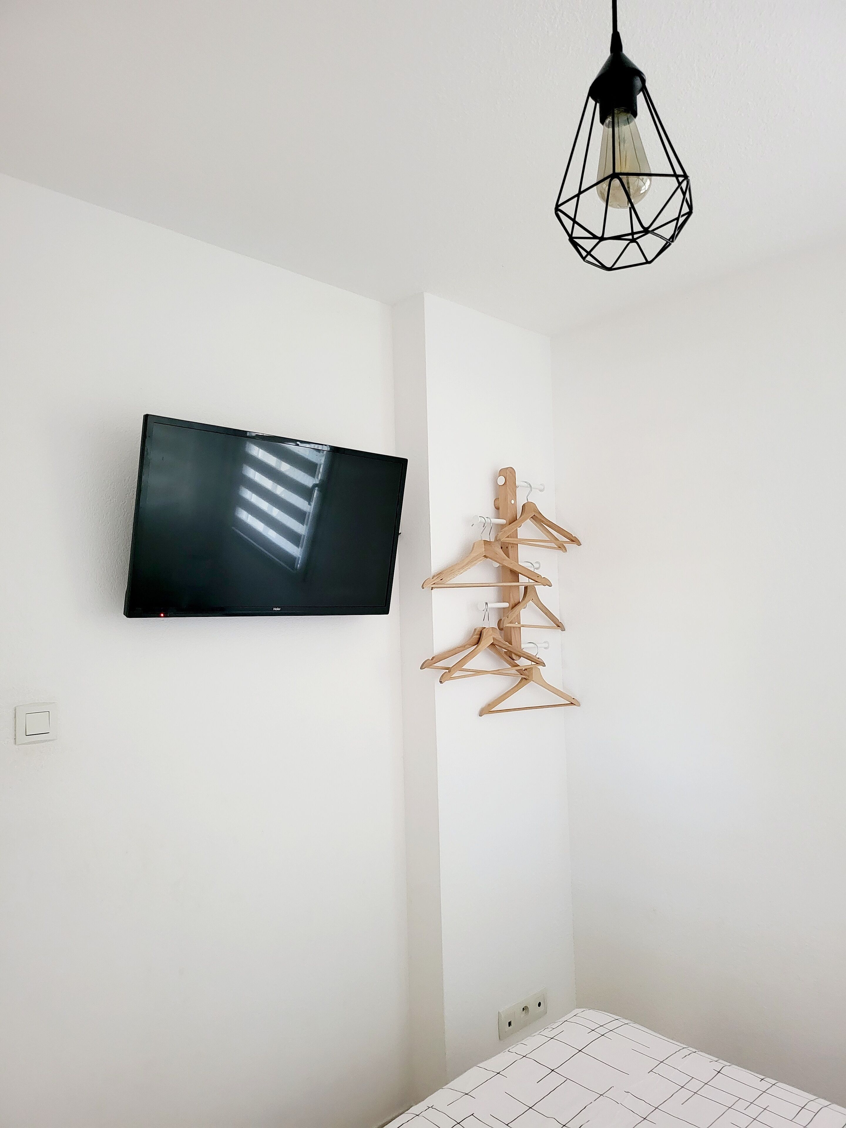 Una stanza con una TV a schermo piatto montata al muro, grucce di legno su un appendiabiti e una lampada a sospensione geometrica.