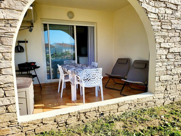 Une terrasse avec une table et des chaises, un barbecue et une vue à travers une porte vitrée.