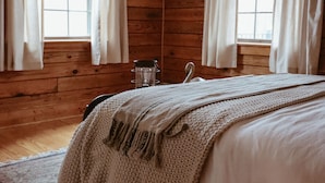 A wooden cabin room with a bed, two windows with curtains, a wooden floor, and a wall with three framed pictures.