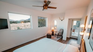 A bedroom with a large bed, a ceiling fan, two windows with views of a field and mountains, a chair, a small table with a lamp, and a door leading to a balcony.