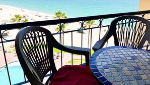 En balkon med to stole og et bord, der ud til en strand med klart turkis vand og en fjerntliggende bysilhuet.