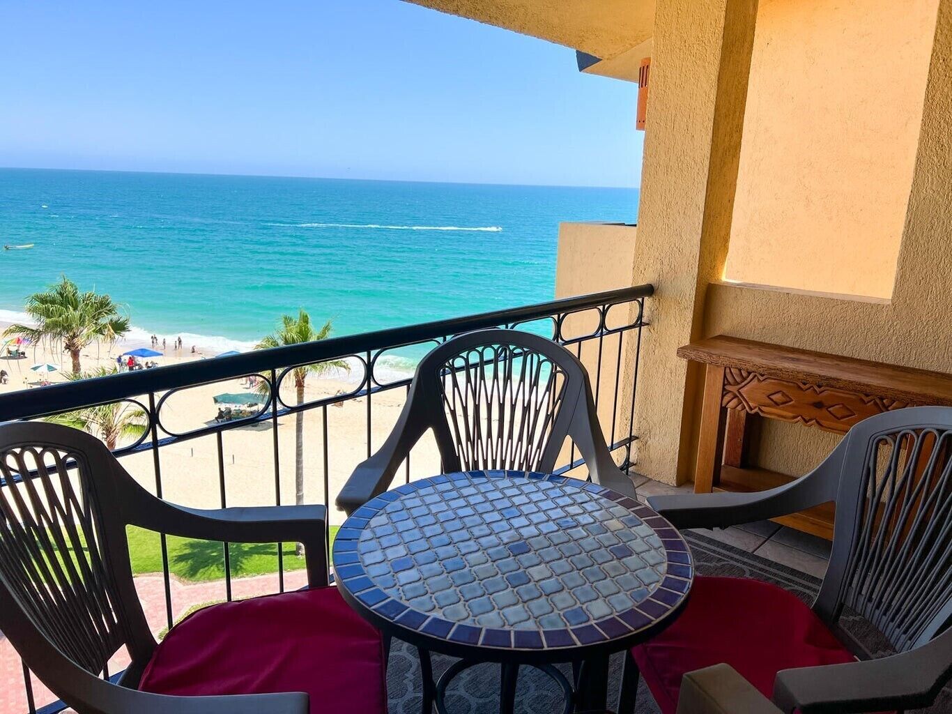 A balcony with a view of the beach and ocean. There are two chairs and a small table with a tiled top.