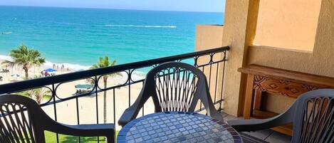 A balcony with a view of the beach and ocean. There are two chairs and a small table with a tiled top.