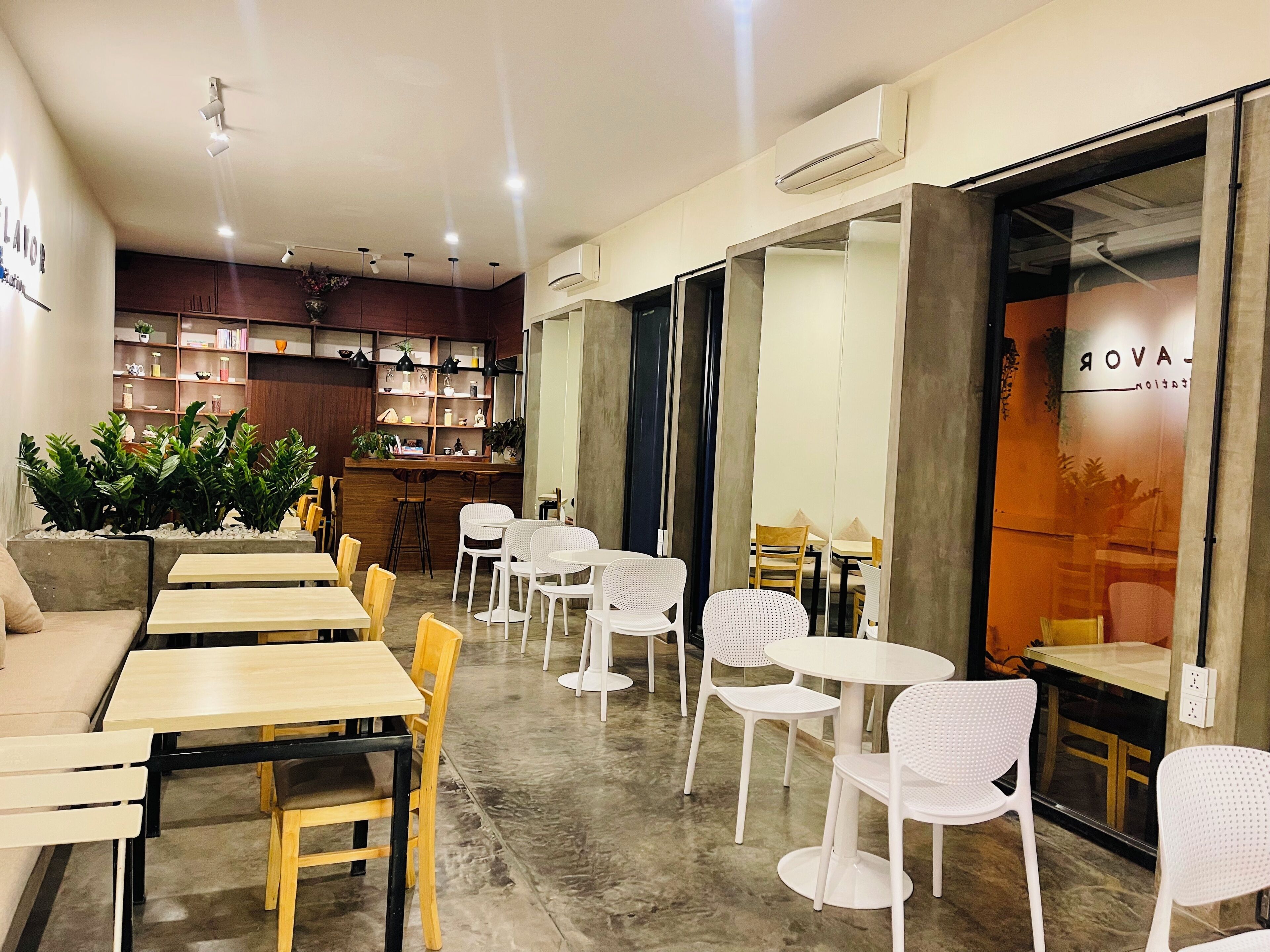 A cafe with multiple seating areas, including tables and chairs, and a bar area with shelves and plants.