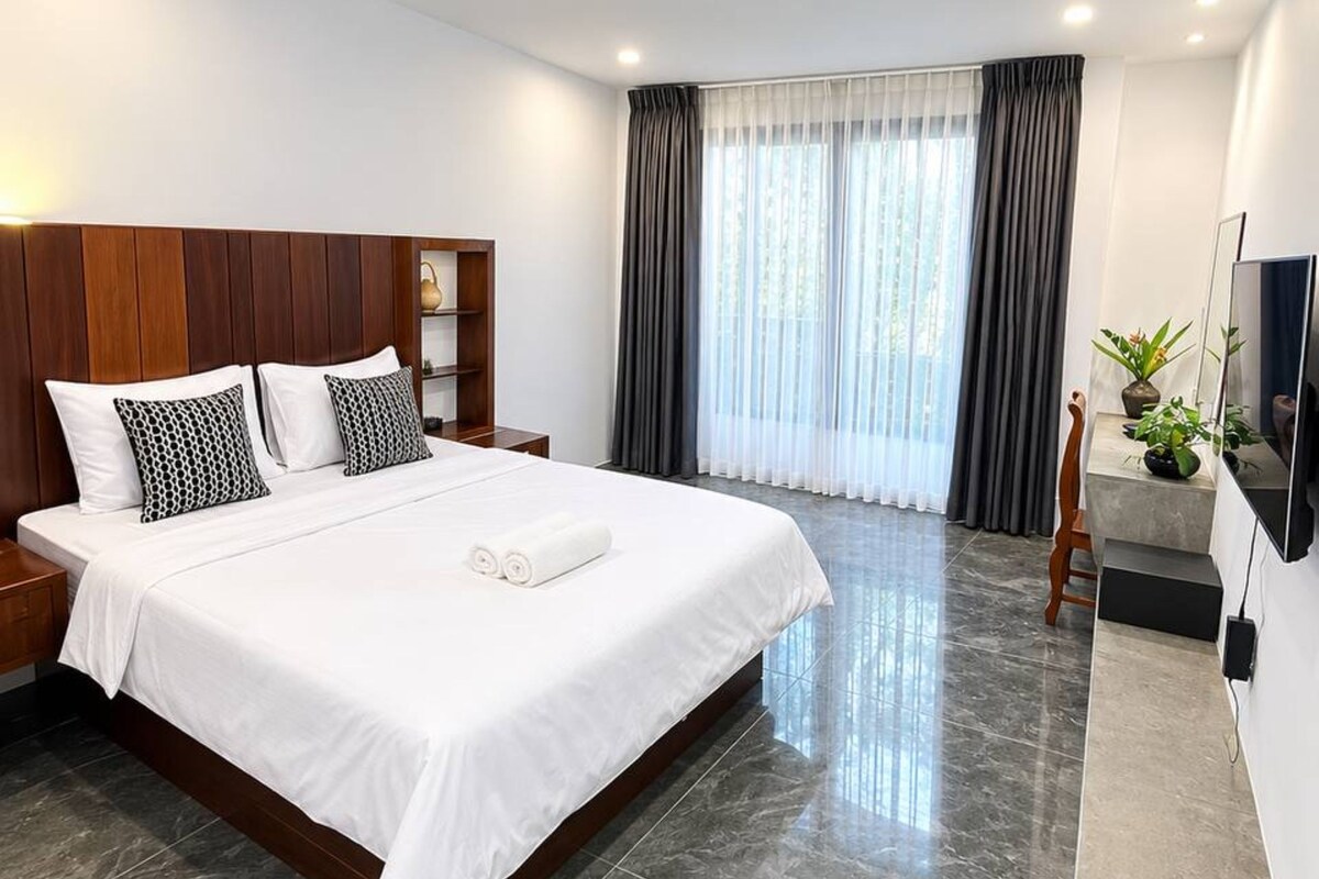 Una habitación de hotel moderna con una cama grande, cabecera de madera, mesita de noche, televisor y un amplio ventanal con cortinas.