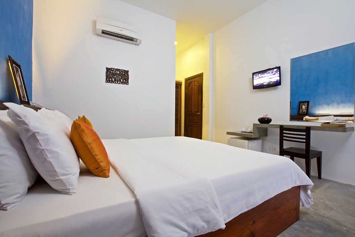 Un dormitorio con cama, ropa de cama blanca, un escritorio con silla, televisor y una puerta que da a otra habitación.