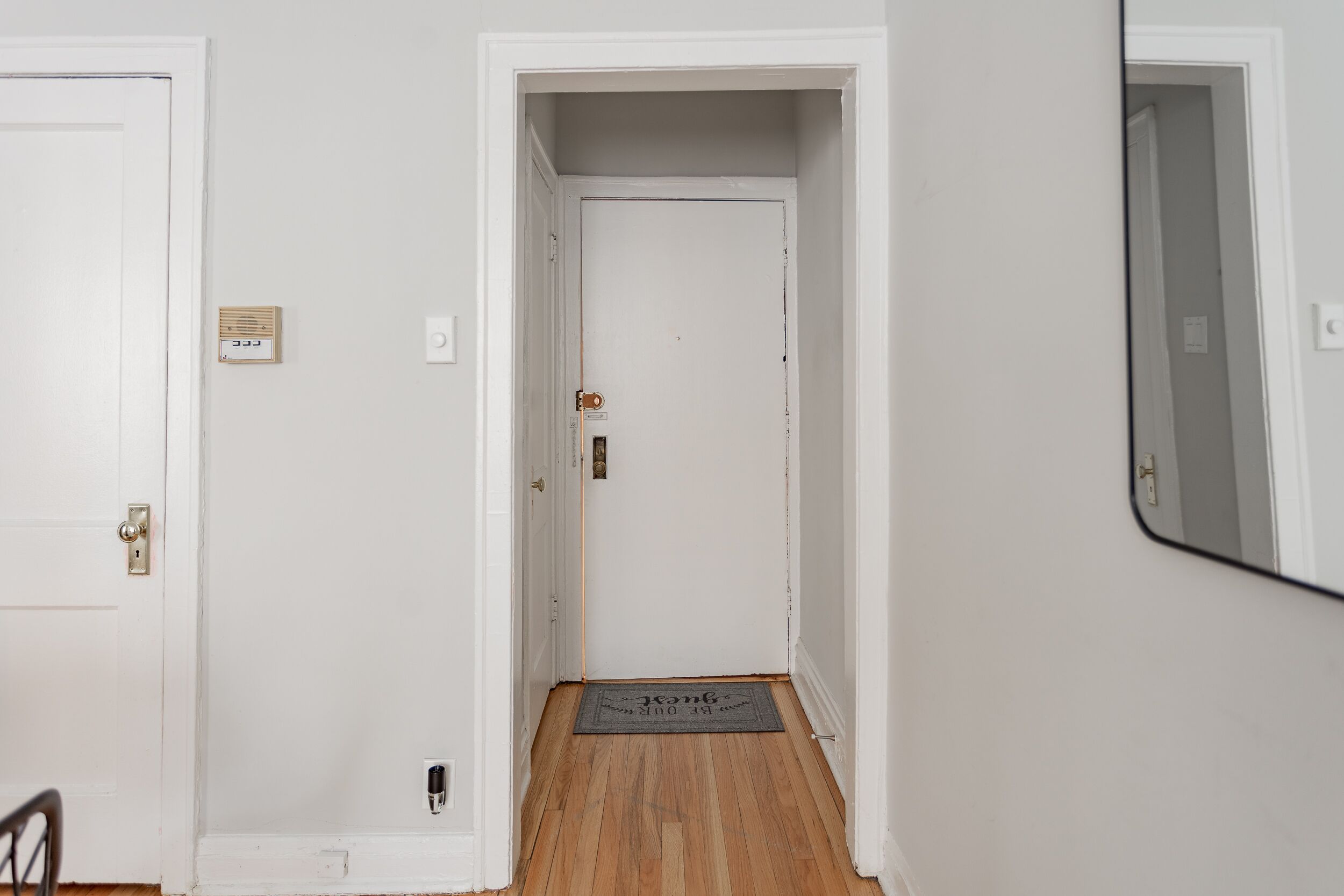 A narrow hallway with a wooden floor, a white door at the end, and a small rug with text.