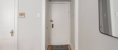 A narrow hallway with a wooden floor, a white door at the end, and a small rug with text.