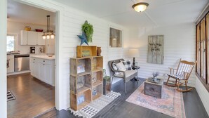 Amplia sala de estar con un sofá, un sillón mecededor y una mesa de centro. Se ve una cocina al fondo con armarios blancos y una lámpara colgante.