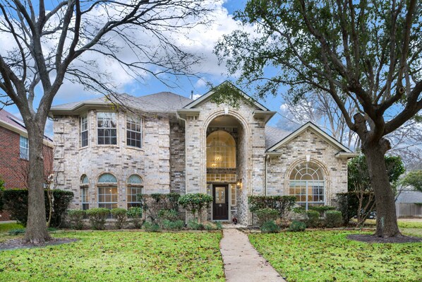 Stately brick building with a central arched entrance and gabled roof, set amidst mature trees and well-manicured grounds.