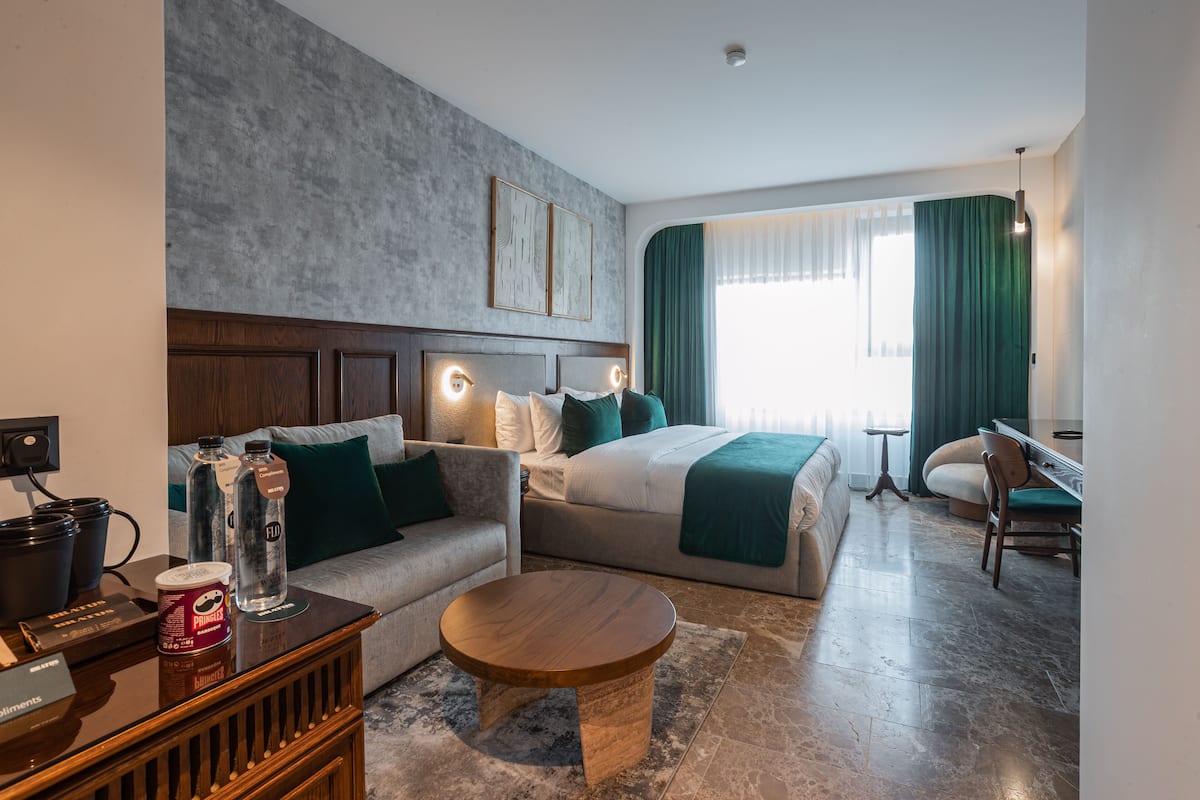A hotel room with a bed, a sofa, a coffee table, a desk, and a chair. There are two paintings on the wall, a window with curtains, and a lamp.