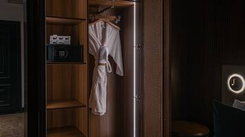 A hotel room with a wooden wardrobe, a robe hanging on the door, a bedside table with a lamp, and a round mirror on the wall.
