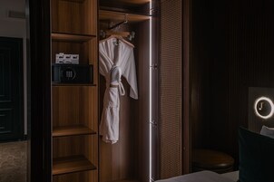 A hotel room with a wooden wardrobe, a robe hanging on the door, a bedside table with a lamp, and a round mirror on the wall.
