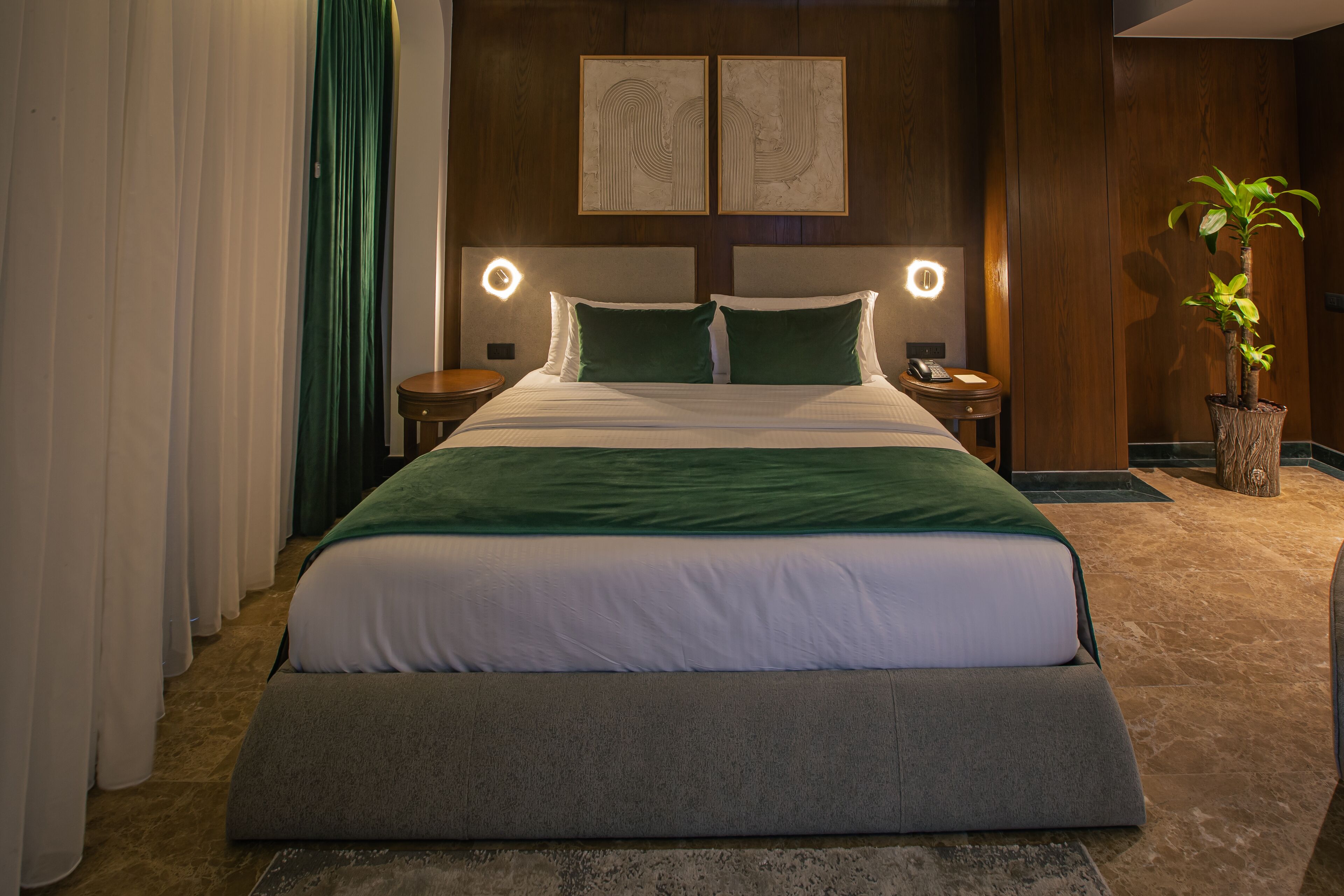 A neatly made bed with a green and white bedspread, two bedside tables with lamps, a potted plant, and two framed artworks on the wall.