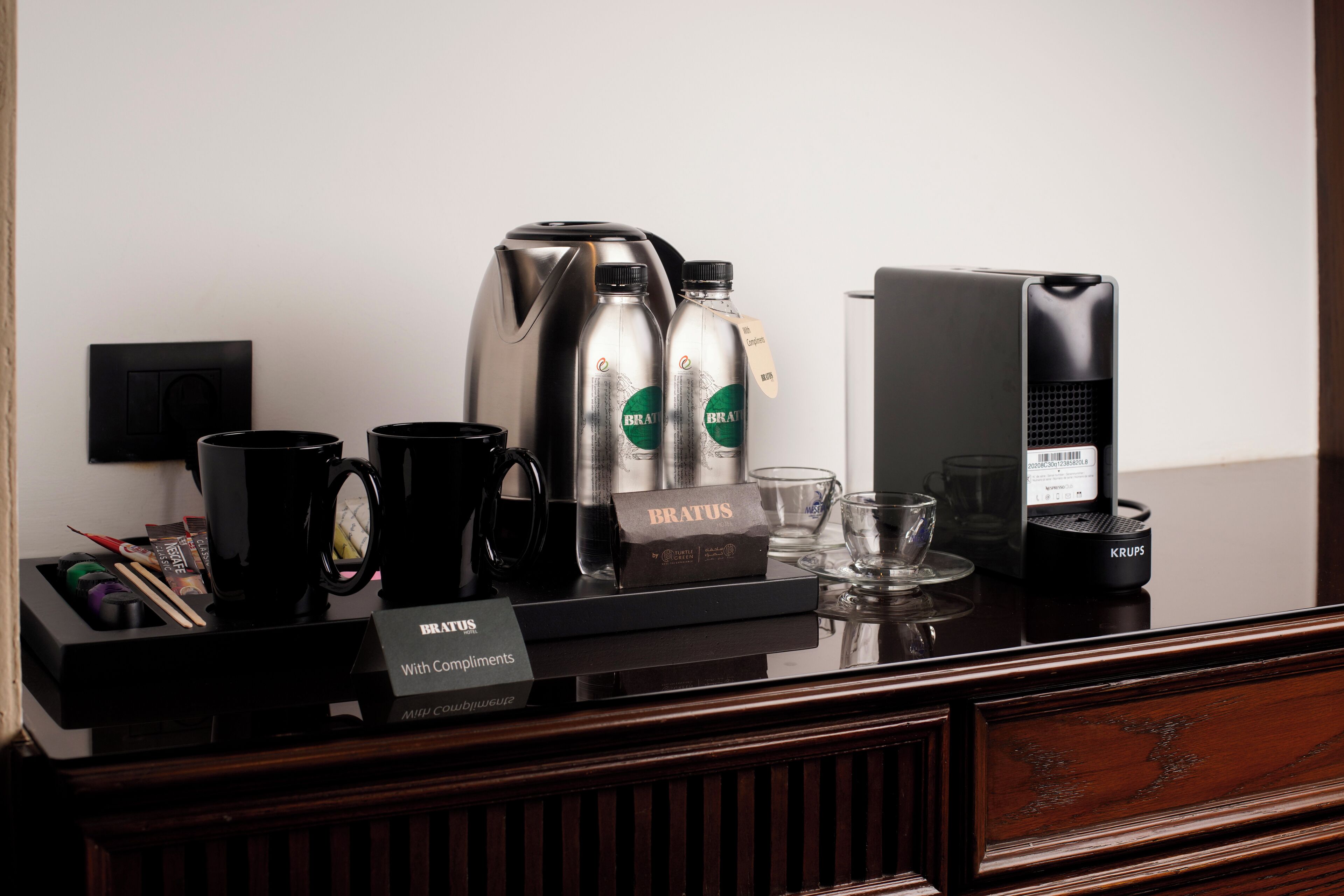 A coffee station with a coffee maker, two bottles of BRATUS, a Krups coffee grinder, and a sign that says "BRATUS With Compliments".