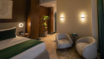 Habitación de hotel con cama, dos sillones, una mesita, una planta e iluminación en la pared.