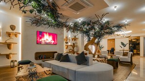 Una sala de estar moderna con un amplio sofá en forma de L, una mesa de centro y un reposapiés verde. Hay un televisor montado en la pared, y un árbol con ramas colgantes es el punto central de la habitación.