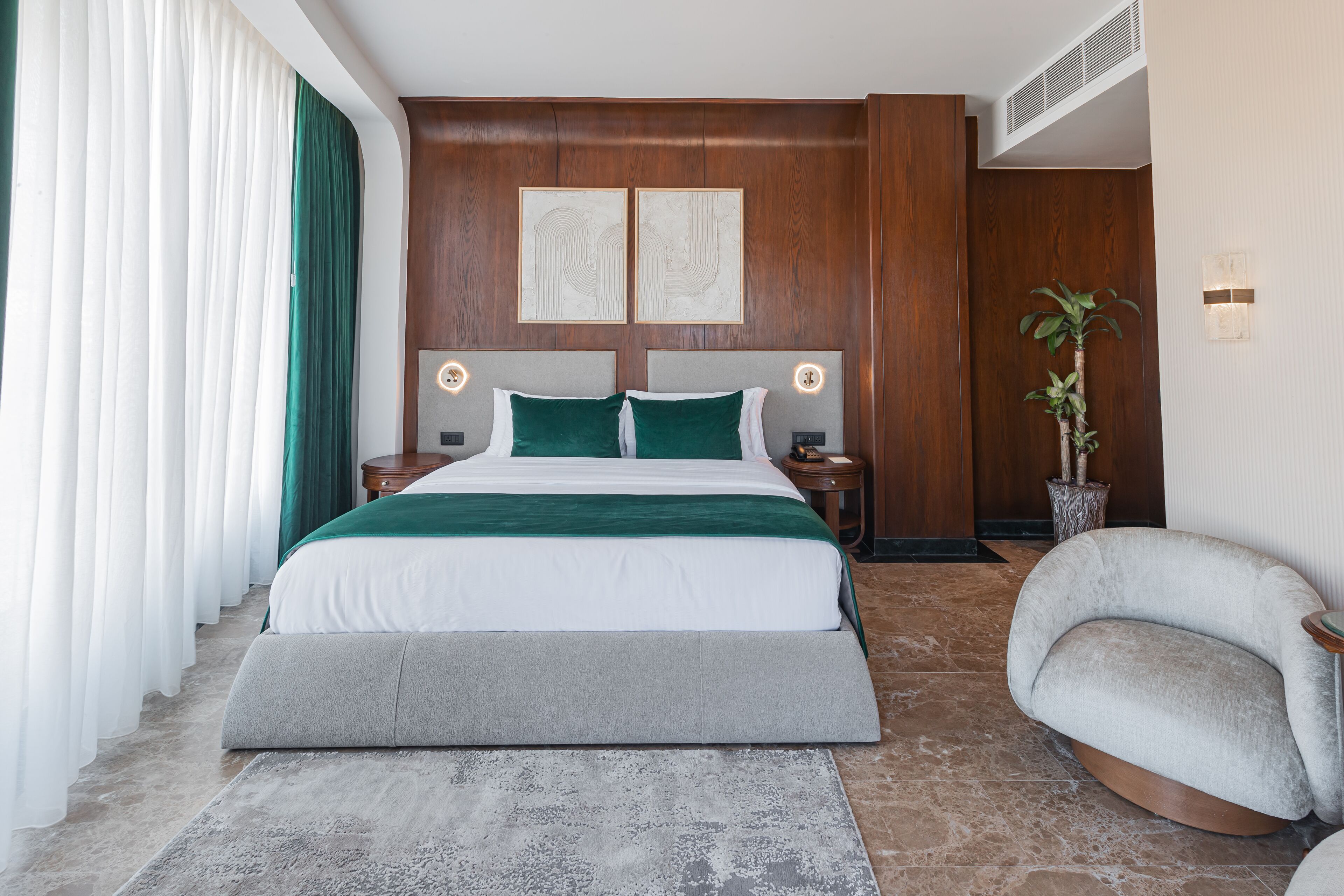 A modern hotel room with a large bed, two bedside tables, a nightstand, a potted plant, and a curved armchair.