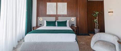 A modern hotel room with a large bed, two bedside tables, a nightstand, a potted plant, and a curved armchair.
