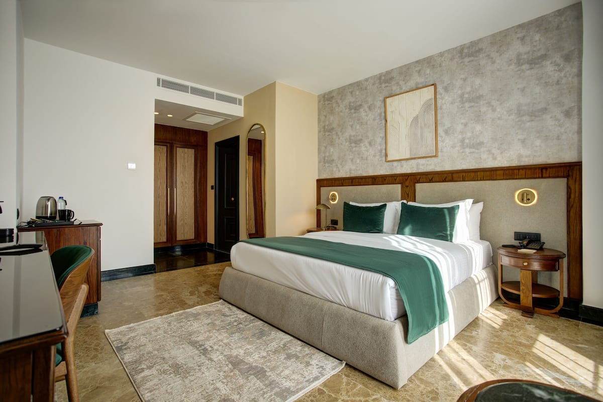 A hotel room with a large bed, a desk with a chair, a small table, and a wall-mounted lamp.