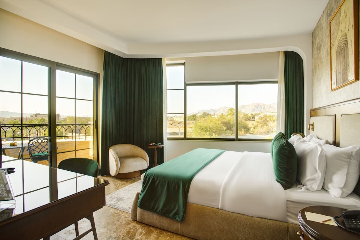 A hotel room with a bed, a desk, a chair, a window with a view of the city, and a balcony with a chair.