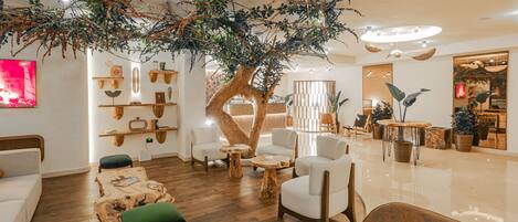 A modern lounge area with wooden flooring, white sofas, and a central tree-like structure.