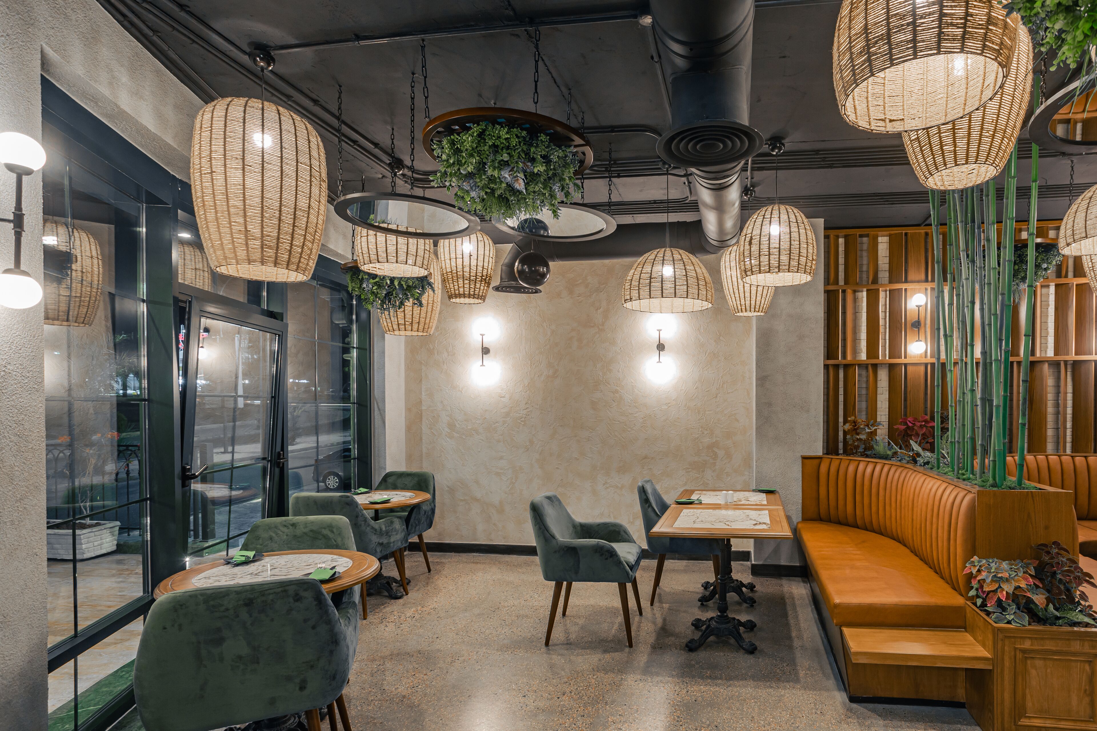 A restaurant interior with round tables, cushioned seating, hanging wicker lamps, and a green plant centerpiece.