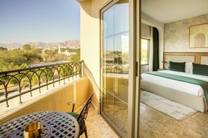 A balcony with a table and chair, a bed with a green bedspread, and a view of mountains and buildings. - Bratus Hotel Aqaba (Aqaba)