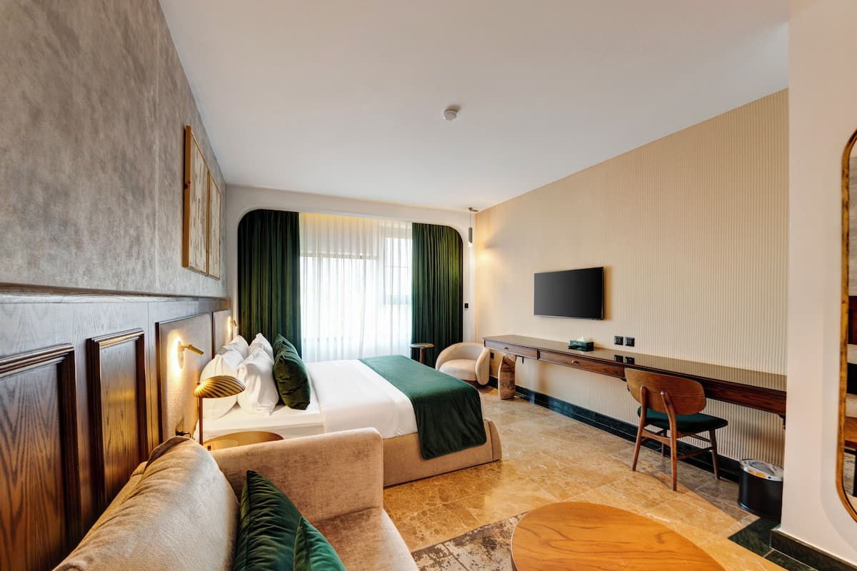 A hotel room with a bed, a sofa, a desk, a chair, a TV, and a window with curtains.