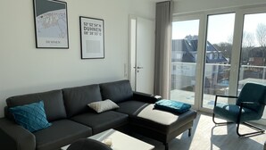 A modern living room with a black leather sofa, a white coffee table, and a blue armchair. There are two framed maps on the wall and a sliding glass door leading to a balcony with a view of houses.