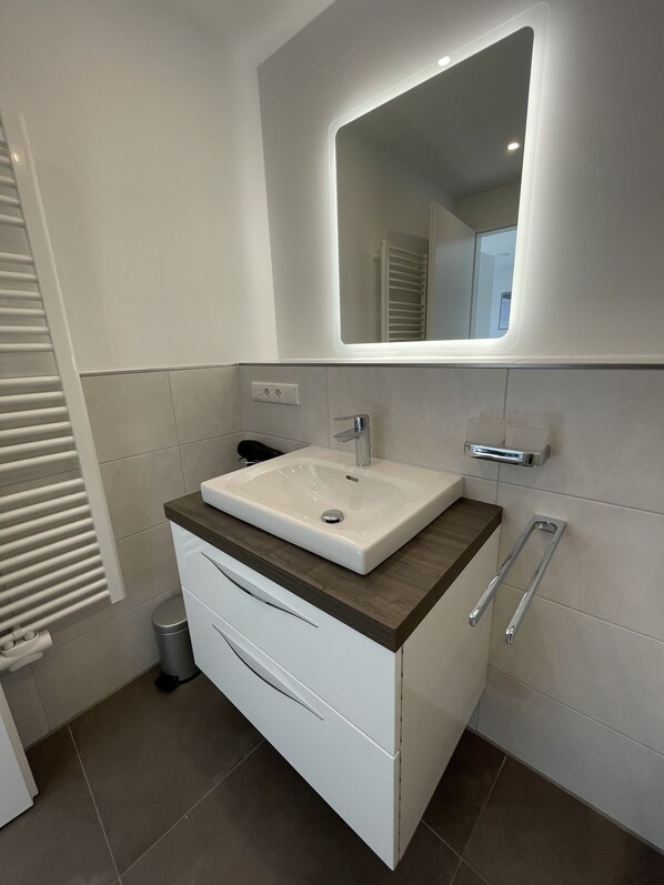 Un baño moderno con un tocador blanco, un lavamanos rectangular, un espejo con iluminación integrada y un perchero para toallas.