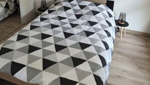 Ein modernes Schlafzimmer mit einer zart gemusterten Bettdecke, einer schwarzen Pendelleuchte, Nachttischen mit Lampen und einem großen Spiegel.