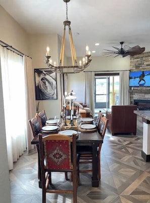 Dining area with a table for six, surrounded by traditional chairs and a chandelier, with a large window and television.