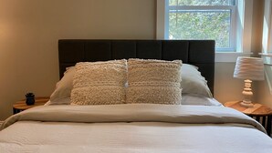 Un llit ben fet amb llençols blancs i coixins decoratius, un panell de capçal fosc, una taula de nit amb una làmpada, i una finestra amb persianes.