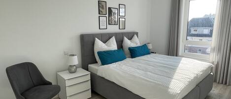 A modern bedroom with a large bed, a bedside table, a chair, and a window with curtains.