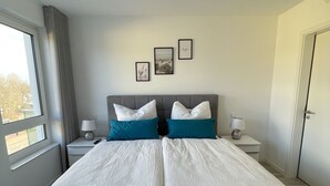 A double bed with grey and white bedding, two bedside tables with lamps, a window with curtains, and three framed pictures on the wall. - Vacation apartment Sunrise 10 with balcony and sea view, Cuxhaven Duhner Allee (Cuxhaven)