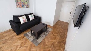 Una sala de estar con un sofá negro, una mesa de centro, un televisor montado en la pared y un suelo de madera con motivo de espiga.