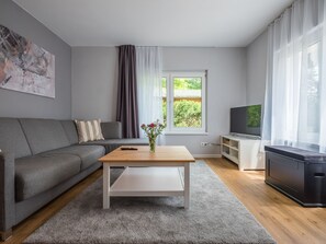 A modern living room with a grey sofa, a wooden coffee table, a TV on a stand, and a window with curtains. - Two bungalows with their own jetty 5 minutes from the lake (Rheinsberg)