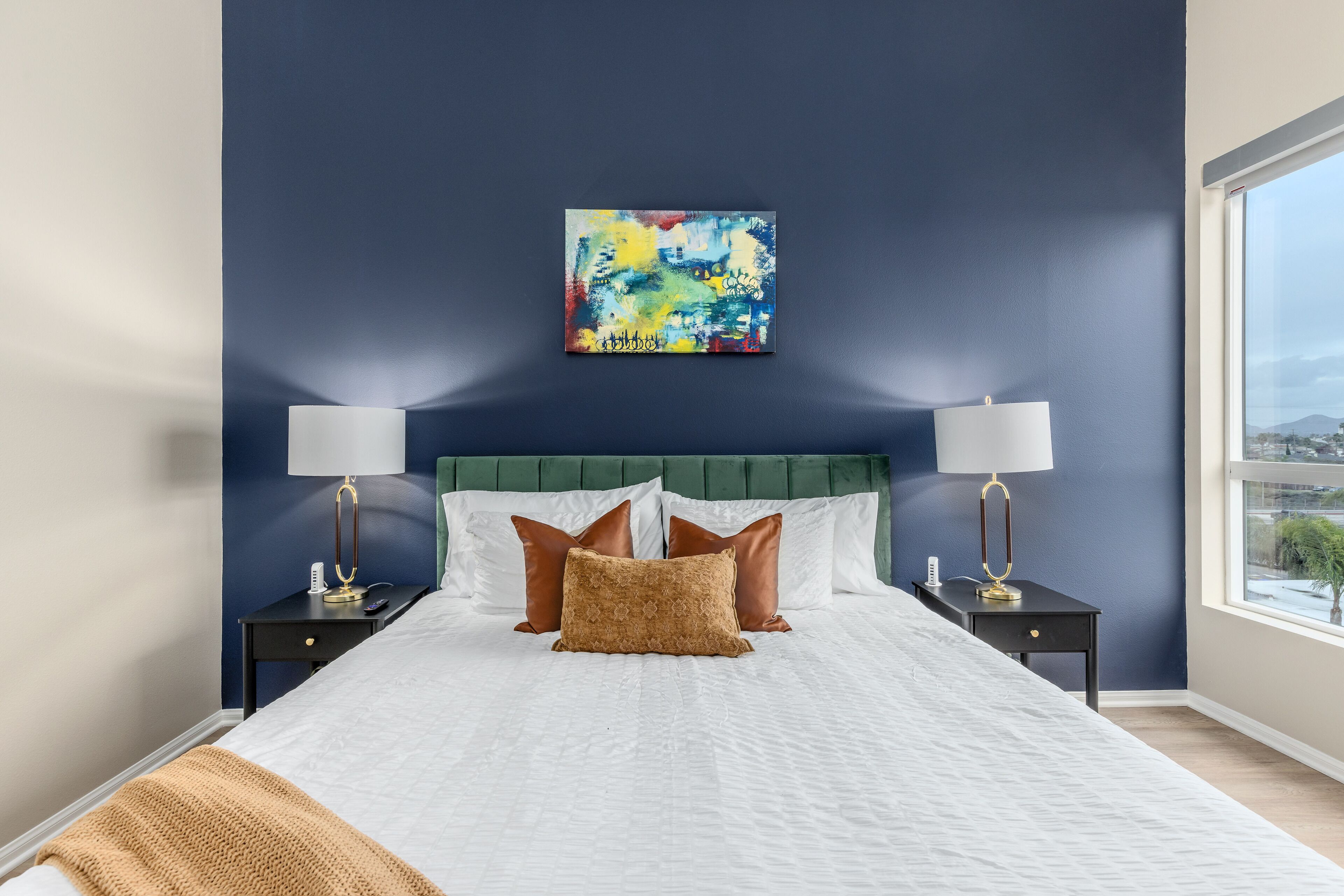 A neatly made bed with white linens, a green headboard, and a colorful abstract painting above it.