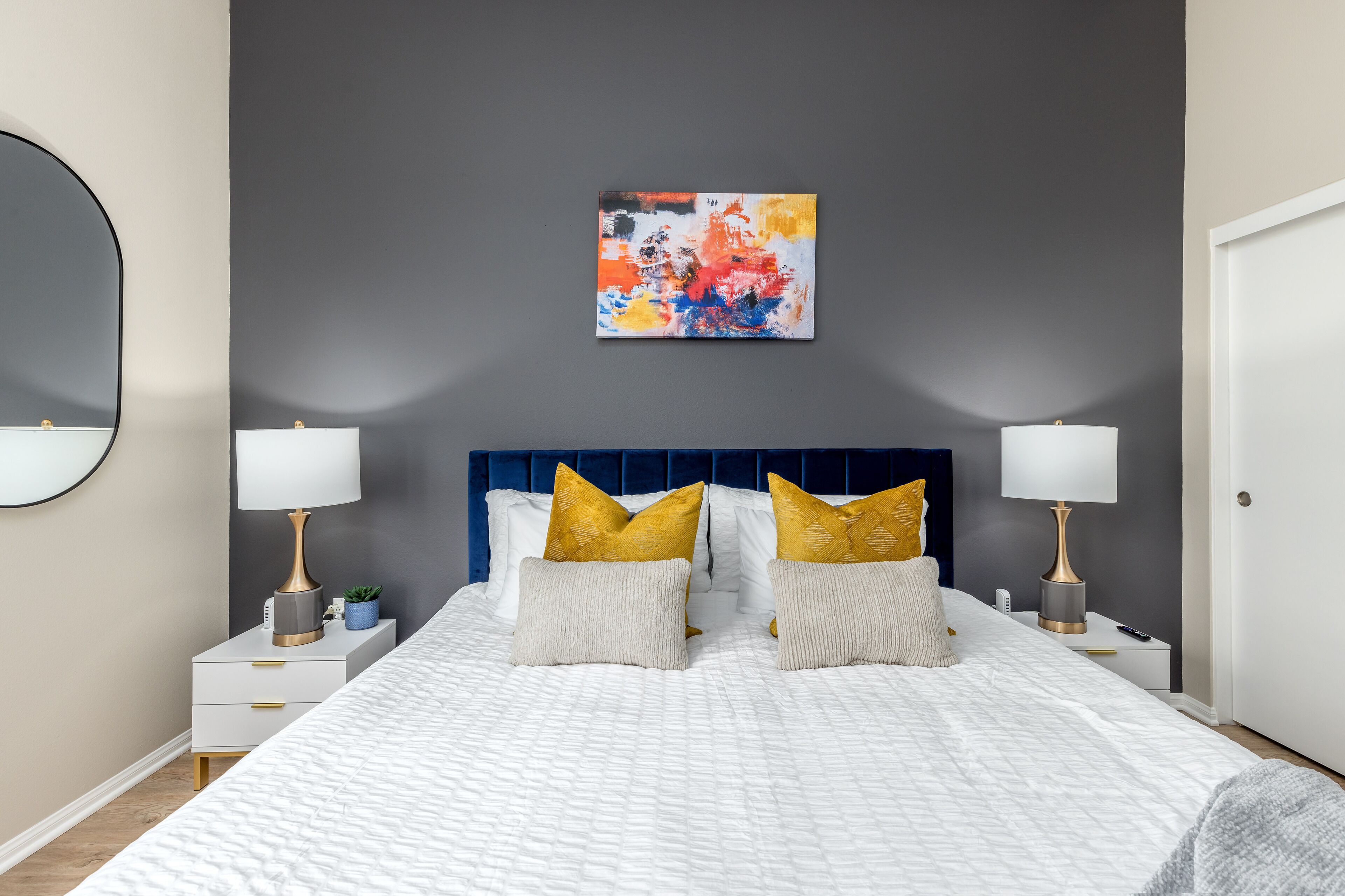 A modern bedroom with a large bed, two bedside tables with lamps, a blue headboard, and a colorful abstract painting on the wall.