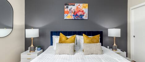 A modern bedroom with a large bed, two bedside tables with lamps, a blue headboard, and a colorful abstract painting on the wall.