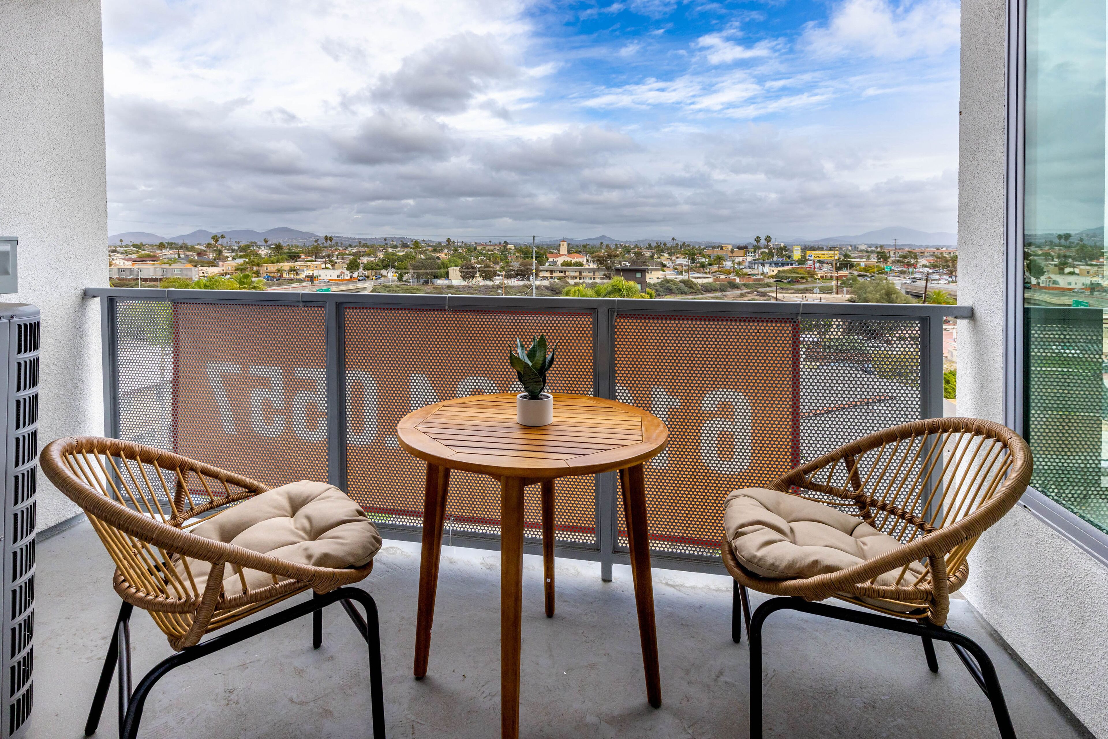 A balcony with two wicker chairs and a small round table, overlooking a cityscape with mountains in the distance.